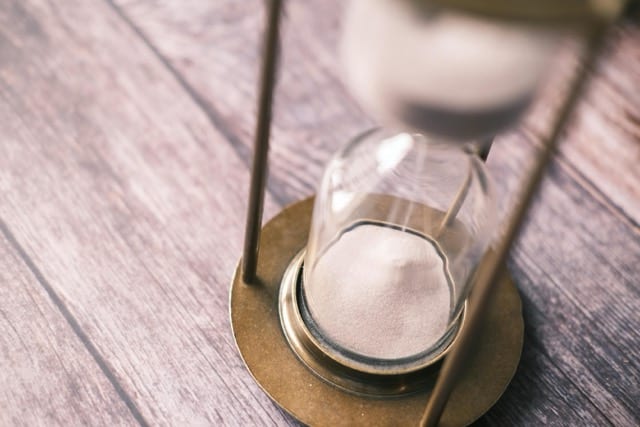 A glass-and-brass hourglass on a dark wooden table. The pale sand has almost entirely fallen to the lower bulb. The narrow neck in the middle is the metaphor for the middle layer—the point where the flow compresses, and through which everything must pass.
