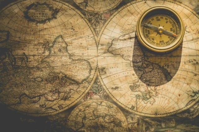 An antique sepia-toned world map in two hemispheres, with a brass compass resting on it. The essay's metaphor: a professional-profile standard is a map, not the territory, and should be read as such.