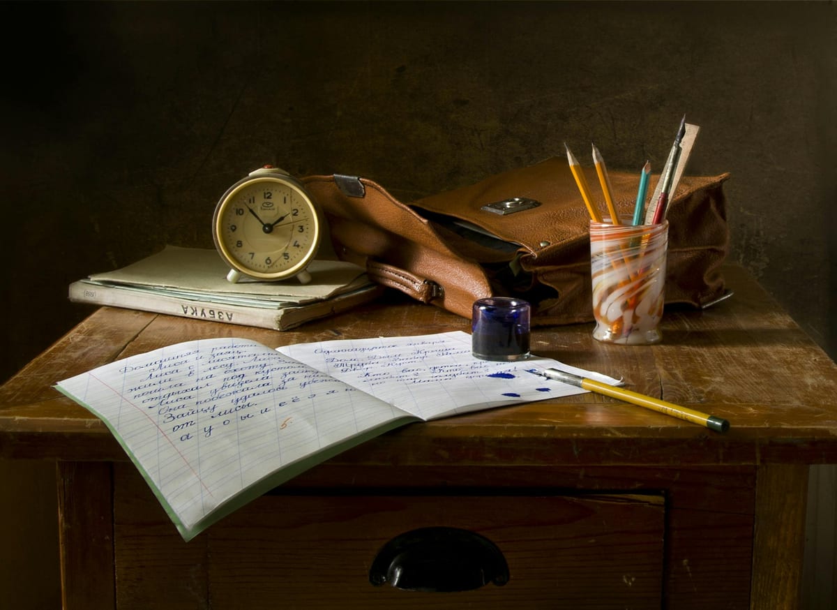 A wooden writing desk with an open notebook full of handwritten script, a blue ink well spilt beside it, a brass alarm clock resting on worn old books. Writing as slow craft, not as form-filling.