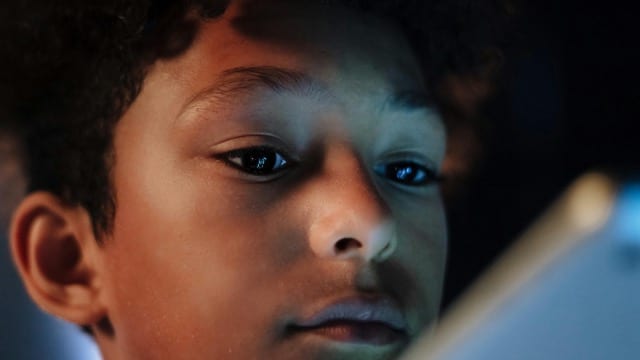A child's face lit by the blue glow of a screen. The eyes are fixed, the attention captured. The skin reflects the colour temperature of the display. Not a dramatic image, an ordinary one. It is what happens in almost every home every evening.