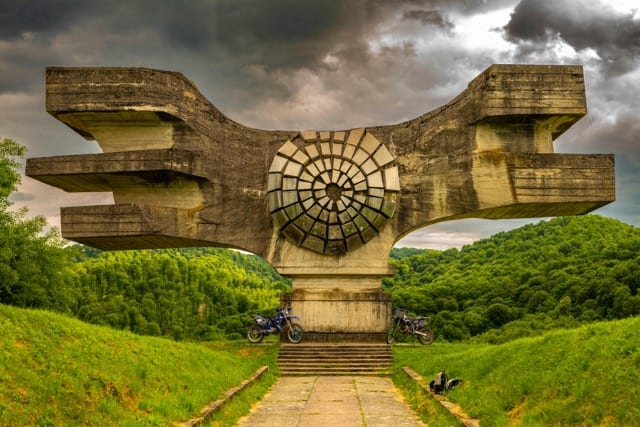 A brutalist concrete monument stands against a stormy sky, two motorcycles parked beneath the arch. A carved disc at the centre, like a stopped clock. The document that describes a system no longer there.