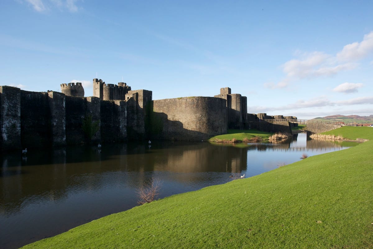 Il castello di Caerphilly, in Galles, visto da oltre il suo fossato: un prato verde in primo piano, l'acqua ferma nel mezzo, le mura grigie sull'altra sponda. Il fossato è la parte più estesa della fortificazione, ed è anche quella che non ha mai combattuto.