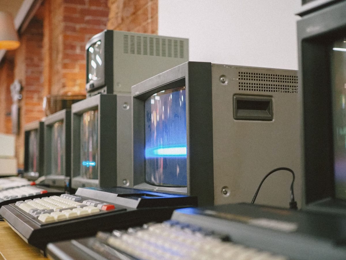 A row of vintage computers lined up on a long wooden table against a brick wall: cathode-ray monitors and mechanical keyboards, one workstation after another. One screen still glows blue. The machines survived; the function that surrounded them didn't.