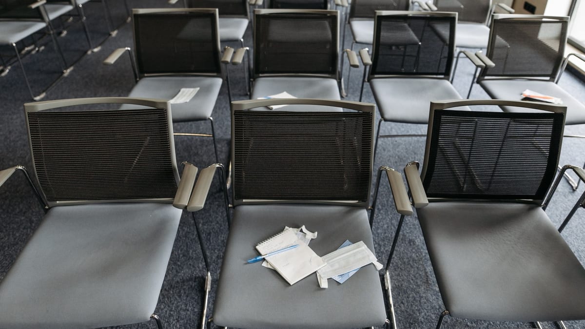 File di sedie identiche in una sala conferenze vuota, viste dall'alto. Su una sola sedia, al centro, restano un blocco per appunti aperto, una penna e alcuni fogli: l'unico posto in cui qualcuno c'era davvero.