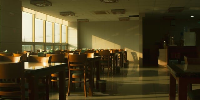 An empty corporate cafeteria in the afternoon, golden light pouring through the windows and casting long shadows on the polished floor. Wooden chairs and tables arranged in waiting, no one seated. The afternoon suspended, before anyone returns.