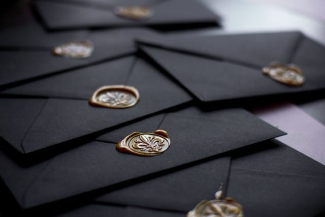 A stack of black envelopes fanned out on a dark surface, each closed with a gold wax seal stamped with a crest. The focus falls on a seal in the foreground, the others fading into the background. The old gesture of guaranteeing the provenance of what is sealed inside.