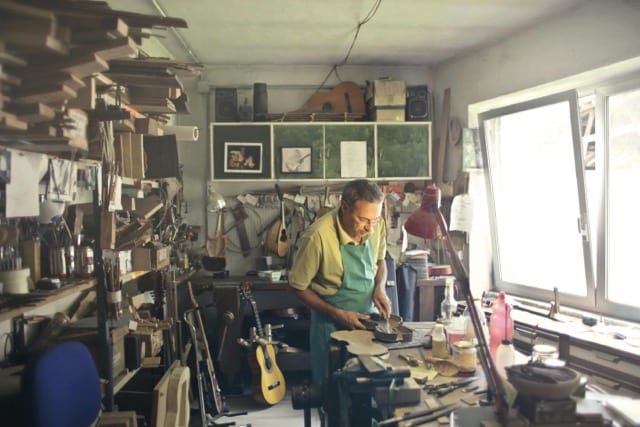 An artisan in his luthier's workshop, surrounded by wood, tools, instruments hanging on the walls. He works on a piece under the light of a desk lamp, in a green apron. The craft built by hand, before the market gives it a name.