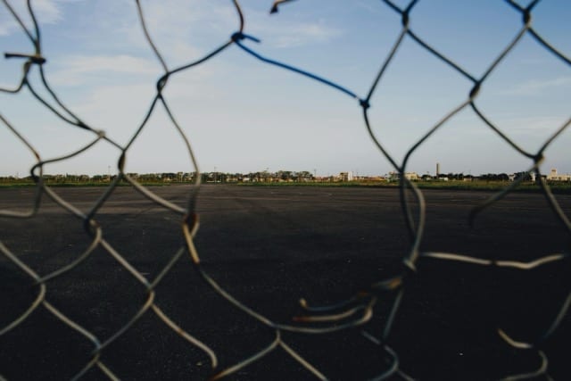 A torn chain-link fence in the foreground, beyond which an empty asphalt expanse opens at dusk. A physical boundary punctured, no longer separating anything. The contract as a perimeter that no longer holds.