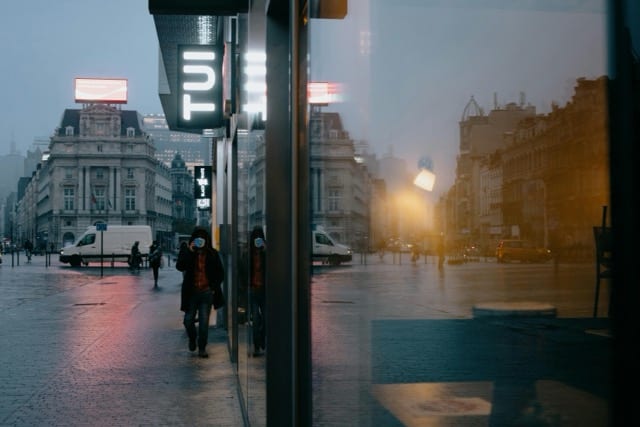A European street at dusk, seen from a glass bus shelter. A figure walks through the orange glow of lit signs and streetlamps. Their image is reflected in the glass beside them, doubled. The spectre is the reflection, the watcher, both at once.