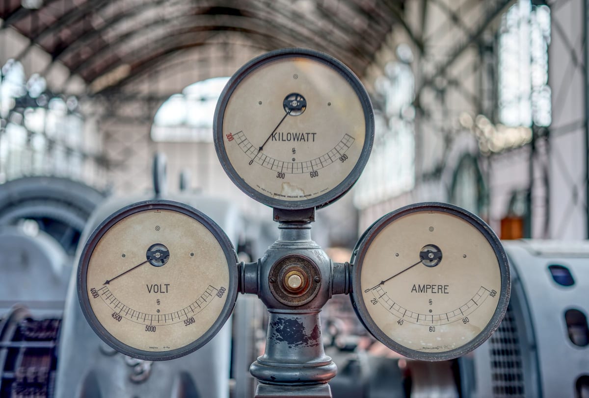 Tre strumenti di misura d'epoca in una sala macchine industriale: un voltmetro, un amperometro e un contatore di kilowatt, montati su una colonna di metallo grigio, con le lancette ferme. Gli strumenti sono perfettamente leggibili; la corrente che dovevano misurare non passa più.