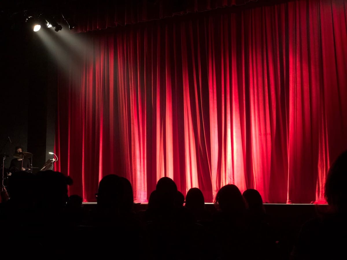 Sipario rosso chiuso su un palcoscenico teatrale, illuminato da un unico faro dall'alto, con le sagome del pubblico già seduto in platea. Nessuna prova generale: quando il sipario si alza, la prima replica è il debutto.