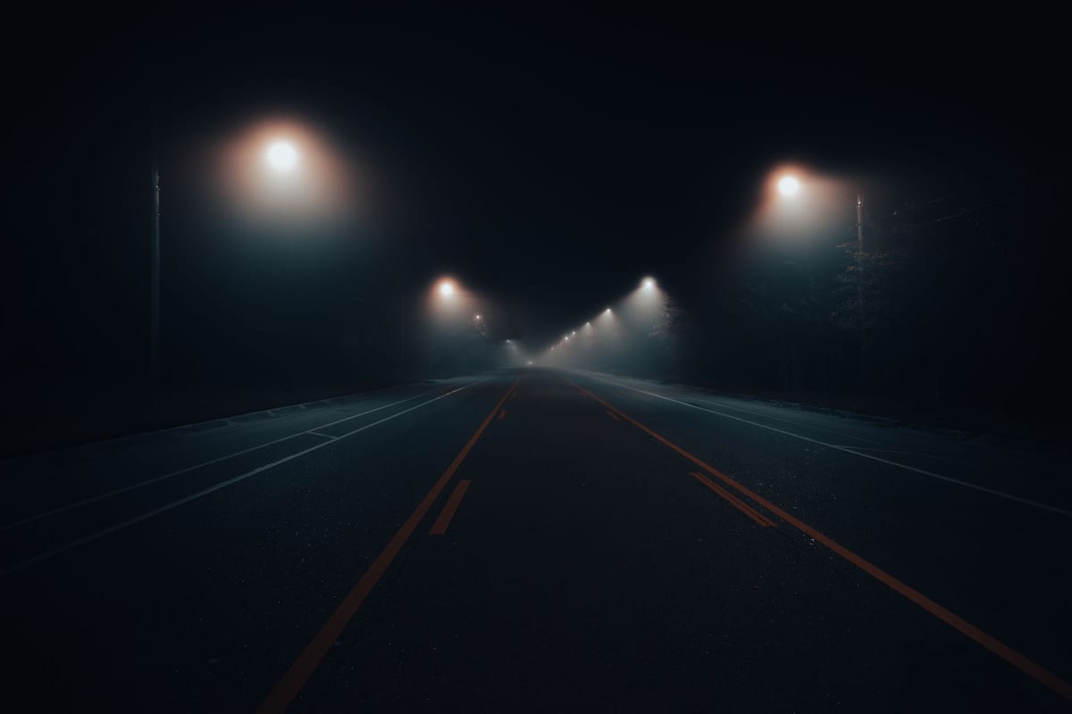 Una strada a due corsie completamente vuota, di notte, nella nebbia. La segnaletica gialla e bianca converge verso un punto di fuga buio, sotto pochi lampioni che si spengono in lontananza. Nessuna auto, nessuna persona.