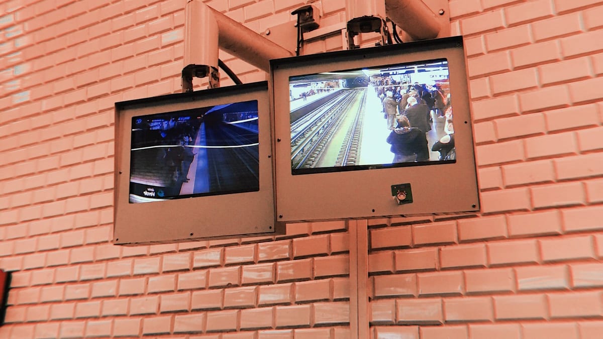 Due monitor di videosorveglianza montati in alto su una parete di mattonelle rosa in una stazione della metropolitana. Uno mostra una banchina affollata di viaggiatori, l'altro una banchina deserta e in penombra. Sopra i monitor sono fissate le telecamere. Nessuno guarda gli schermi.